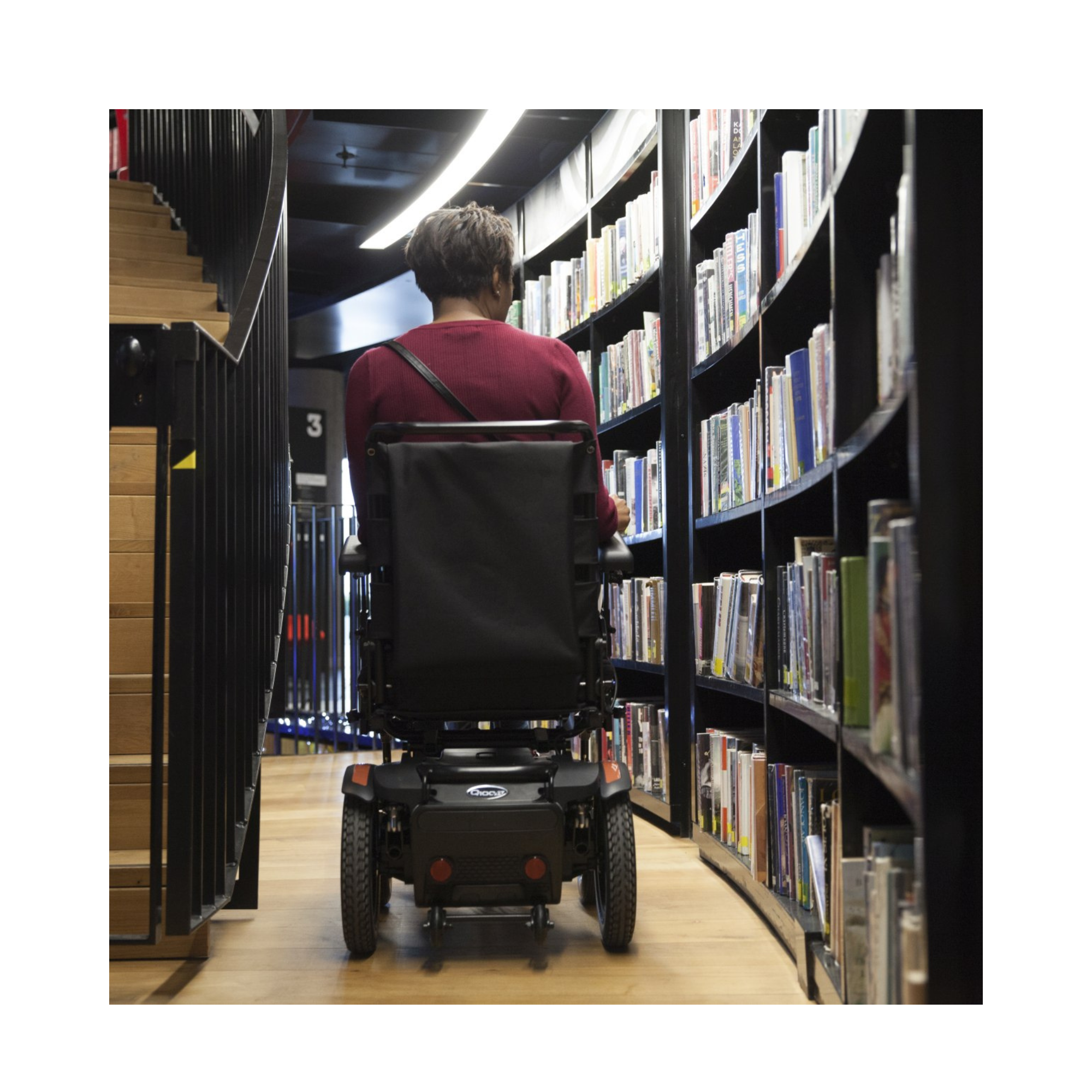 Mujer se desplaza en silla de ruedas eléctrica por un pasillo estrecho entre estanterías de una biblioteca
